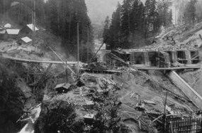 Historic black-and-white photograph of Elwha Dam under construction in a forested canyon